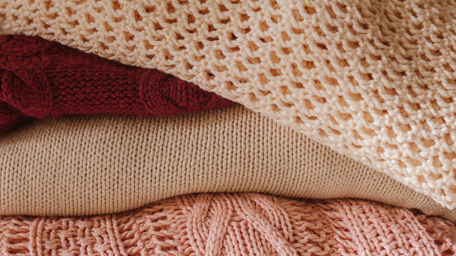A stack of folded knitted wool and cashmere jumpers in cream, beige, burgundy, and pink.