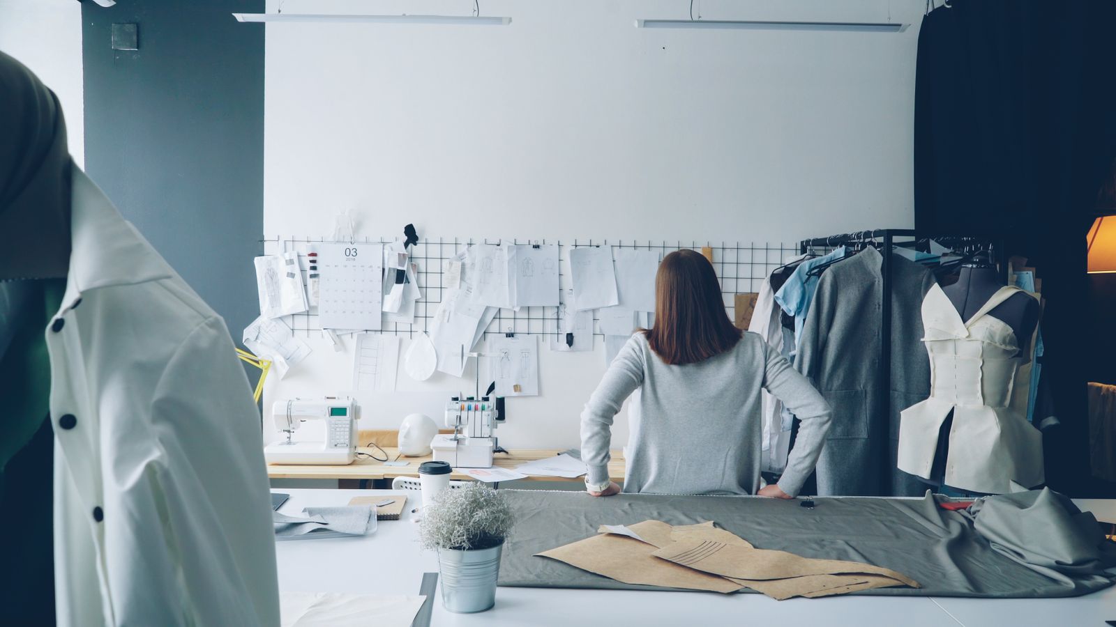 Rear view of a person standing at a cutting table in a fashion design studio beside a mannequin and paper patterns.