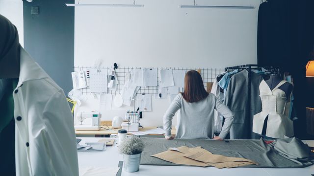 Rear view of a person standing at a cutting table in a fashion design studio beside a mannequin and paper patterns.