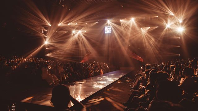 An illuminated fashion runway viewed from the audience with dramatic stage spotlights and silhouettes of spectators.