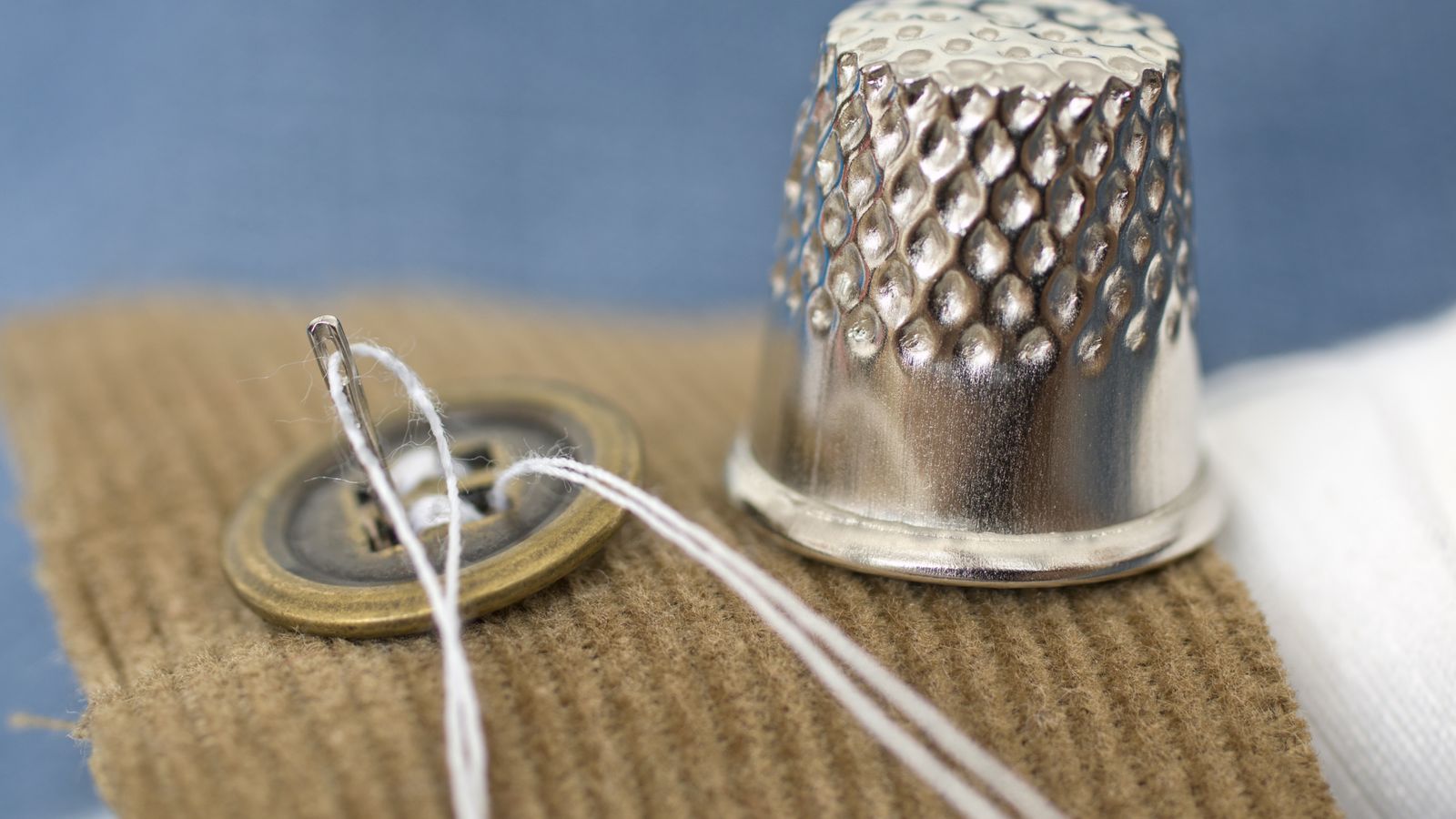 Close-up of a button being sewn onto fabric with needle, white thread, and a metal thimble nearby