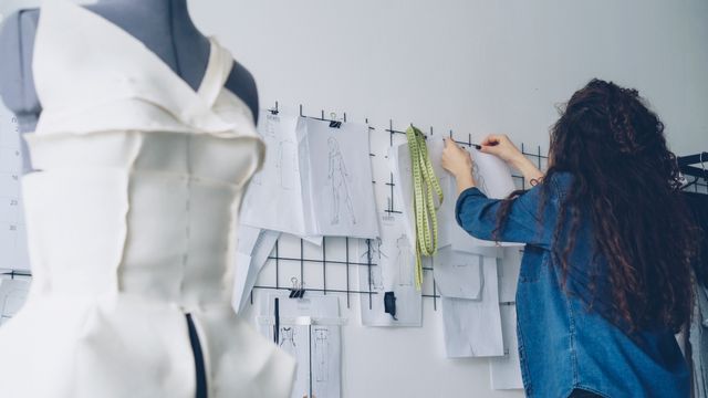 A tailor's mannequin draped in structured white fabric beside a designer working at a mood board with fashion sketches.