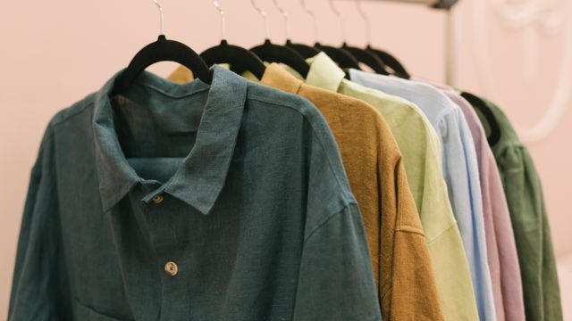 A row of collared linen shirts in green, mustard, sage, and blue hanging on black clothes hangers.