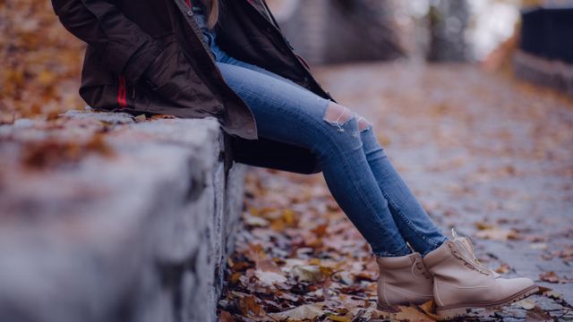 A person sitting on a stone ledge wearing blue denim jeans, an autumn jacket, and ankle boots amongst fallen leaves.