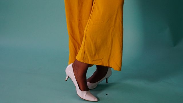 A person wearing wide-leg mustard yellow trousers paired with pointed white kitten heels, against a teal background.