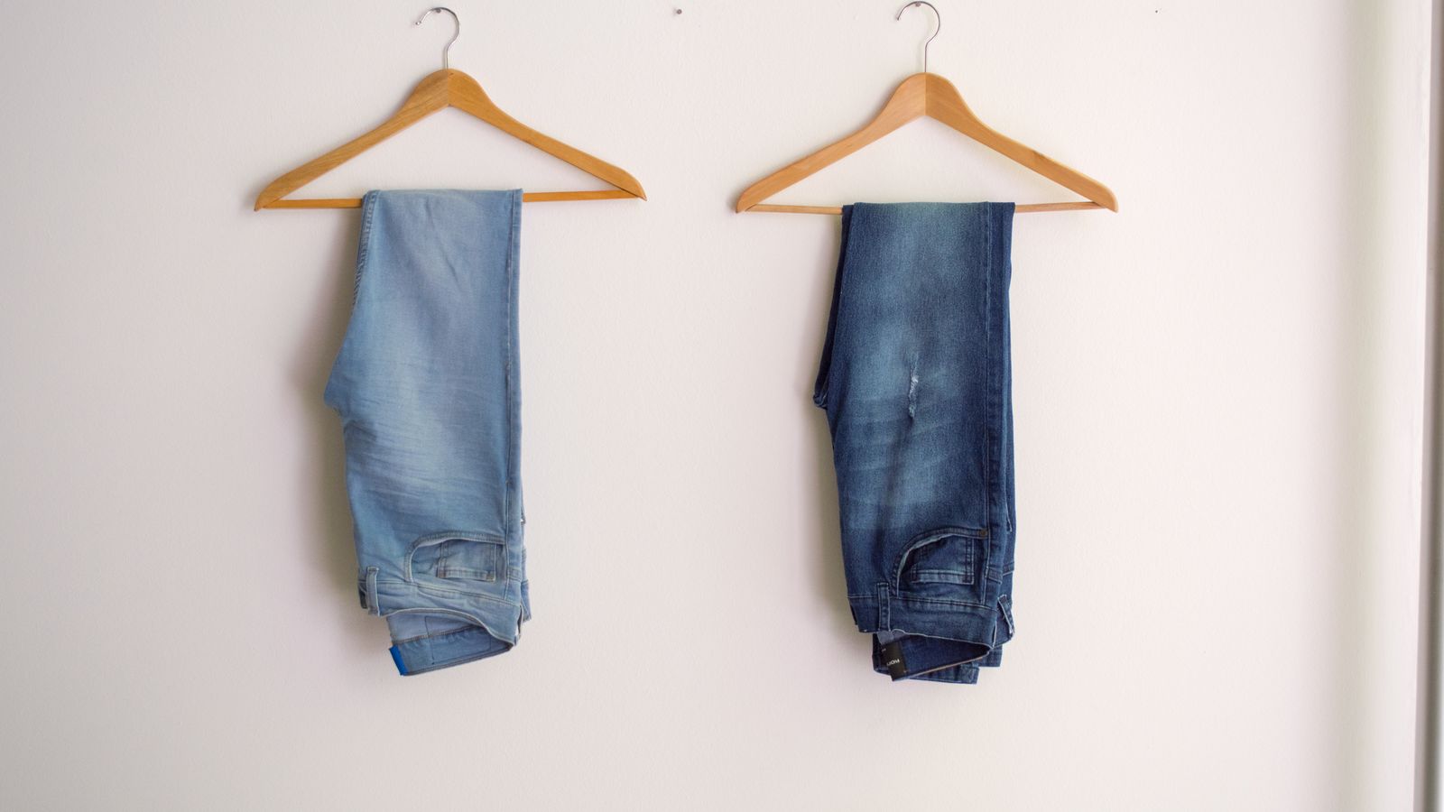 Two pairs of denim jeans hanging on wooden clothes hangers against a white wall.