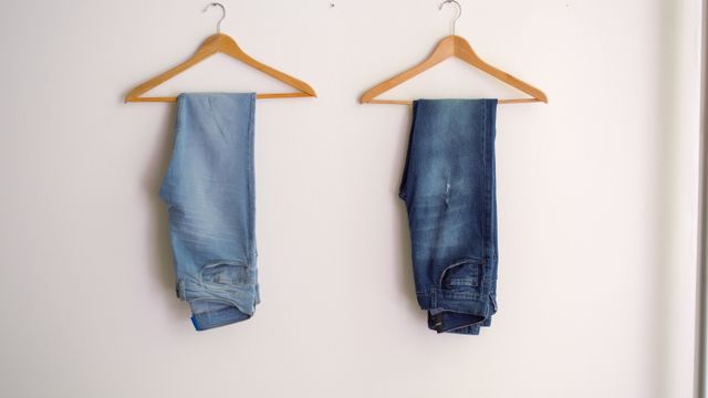 Two pairs of denim jeans hanging on wooden clothes hangers against a white wall.